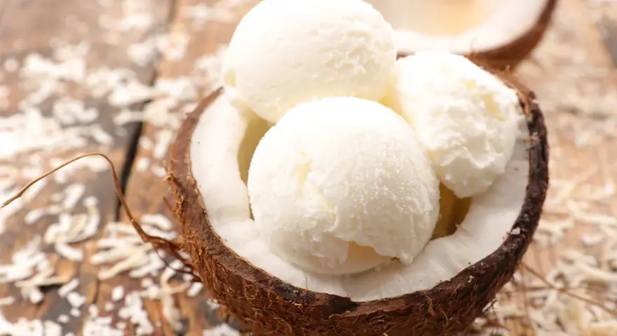 Cremiges Kokosnuss Eis in einer halbierten Kokosnuss, umgeben von Kokosraspeln auf Holz. Ein tropischer Genuss!