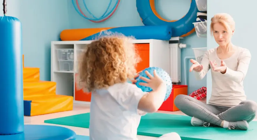 Ergotherapie für Kinder: Eine Therapeutin leitet ein Kind mit lockigem Haar an, das einen blauen Ball hält, in einem bunten Raum voller Spielgeräte.
