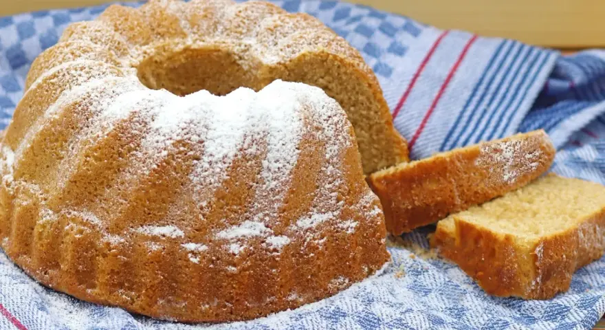 Puszysta babka z cukrem pudrem, obok niej kromki ciasta, na niebieskim ręczniku.