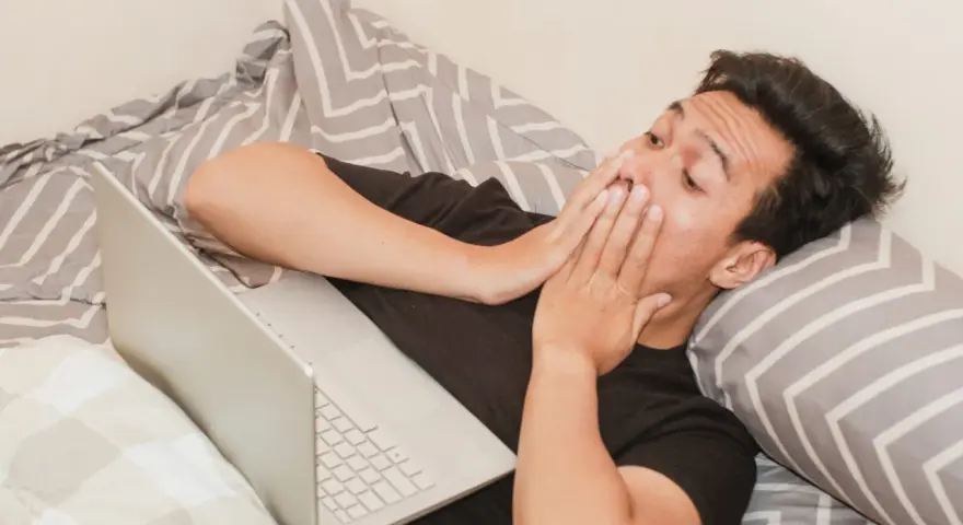 Mężczyzna w łóżku z laptopem, zakrywający usta dłońmi, może symbolizować walkę z uzależnieniem od pornografii.
