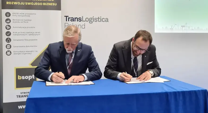Dwóch mężczyzn w garniturach podpisuje dokumenty. Obok baner z napisem TransLogistica Poland, sugerujący umowę w branży gpw logistics.