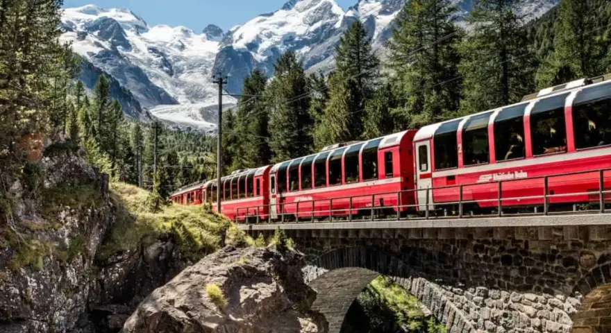 A red train travels across a stone bridge, winding its way from Chur to Tirano through a stunning alpine landscape with snow-capped mountains.