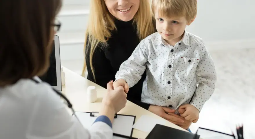 Mama z synkiem u lekarza. Chłopiec ściska dłoń lekarce, a mama uśmiecha się. W tle widać tablet i białą buteleczkę, jakby z lekiem. To ważny moment, być może wpis do książeczki zdrowia.