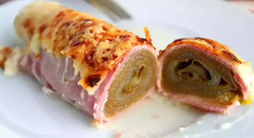 Endive au jambon gratinée, enroulée et coupée en deux, servie sur une assiette blanche avec une sauce crémeuse.