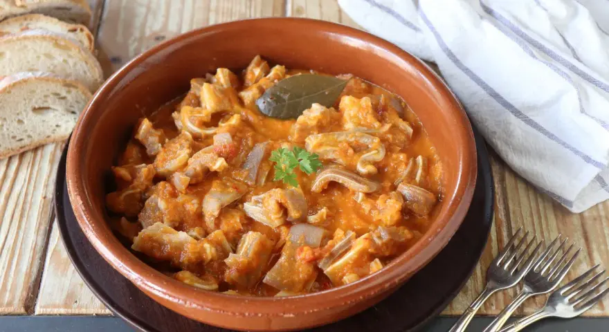 Guiso casero de oreja de cerdo en salsa de tomate, servido en cazuela de barro, acompañado de pan rústico y tenedores.