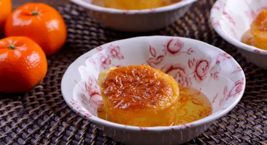 Un esponjoso baba al ron bañado en almíbar, adornado con ralladura de naranja, en un cuenco floral. Cerca, mandarinas frescas.