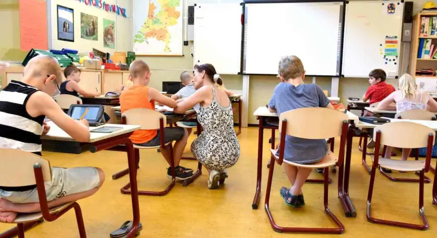 Dzieci siedzą w klasie, ucząc się. Nauczycielka pochyla się nad jednym z uczniów. Ile jest klas w Anglii? Tego nie wiemy, ale ta lekcja wygląda na interesującą.