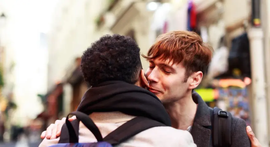 Un hombre joven con cabello rojizo susurra "bonjour" a su pareja, un momento íntimo en una calle parisina.