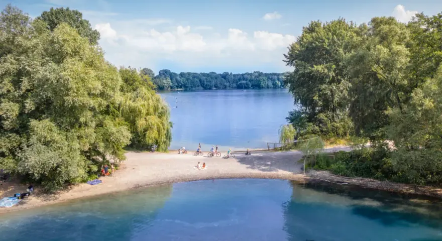 Sommerliche Idylle am Strandbad Wedau: Menschen genießen den Tag am Seeufer, einige radeln, andere entspannen im Sand.