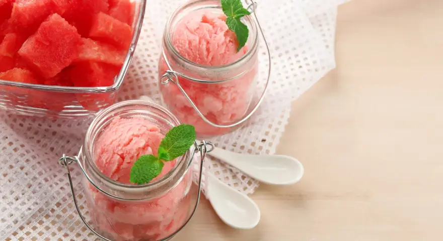 Erfrischendes Wassermelonen-Eis in Gläsern, garniert mit Minze. Daneben ein Schälchen mit frischen Wassermelonenstücken.
