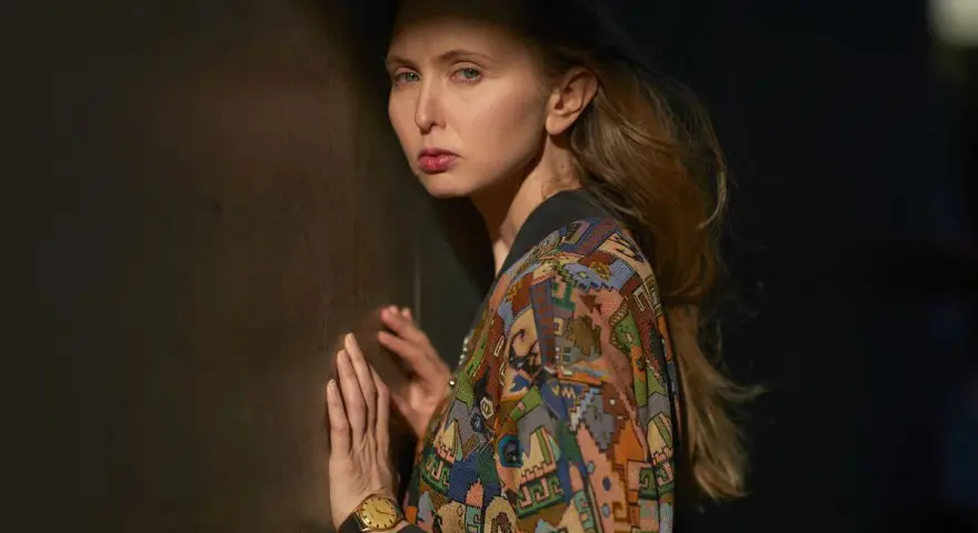 Justyna Wasilewska w kolorowej kurtce opiera się o ciemną ścianę, jej twarz oświetla cień.