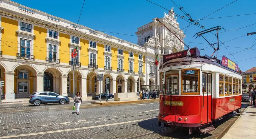 Czerwony tramwaj w Lizbonie, obok żółtego budynku z arkadami. Czy do Portugalii trzeba paszport? Nie, wystarczy dowód osobisty.