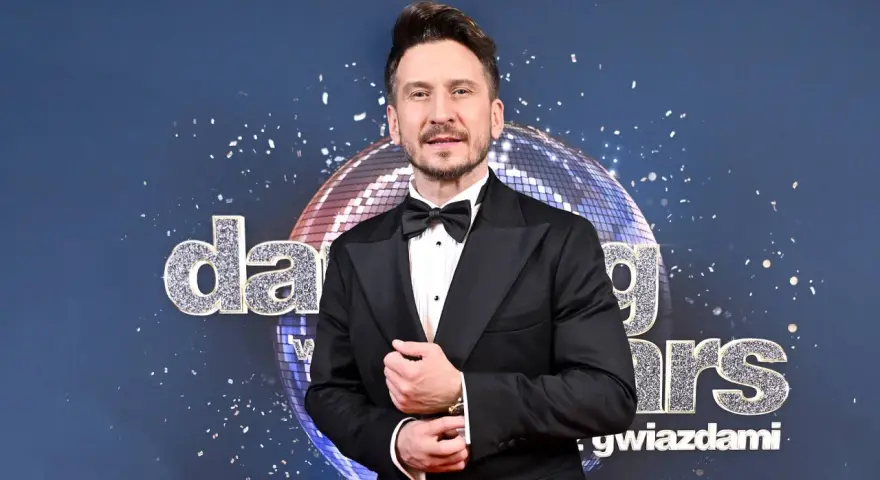 Tomasz Barański w czarnym smokingu pozuje na tle błyszczącej kuli dyskotekowej i napisu "Dancing with the Stars".