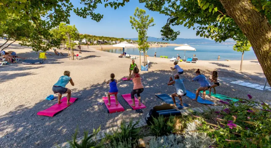 Grupa osób ćwiczy jogę na plaży w Omisalj. W tle widać morze i kemping.