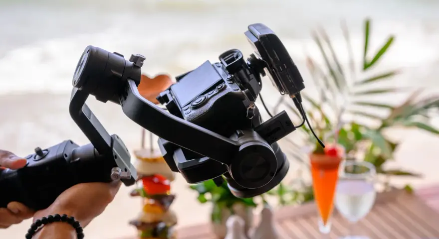 Un gimbal stabilizza una fotocamera per riprese in stile mezzo busto cinema, con un drink e cibo in spiaggia sullo sfondo.