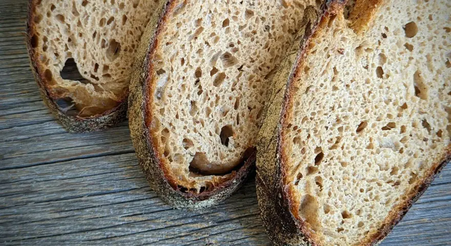 Drei Scheiben rustikales Sauerteigbrot liegen auf Holz. Der Sauerteig ist zu sauer, aber die Kruste ist perfekt gebräunt.