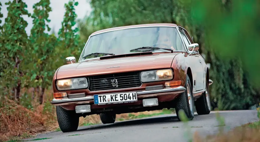 Klasyczny brązowy Peugeot 504 Coupe jedzie po krętej drodze wśród zieleni.