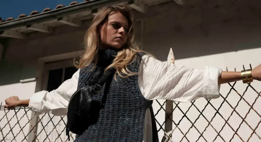 Une jeune femme porte un pull sans manche en maille bleue sur une chemise blanche, un sac banane noir et un bracelet doré. Elle s'appuie sur une clôture rouillée.