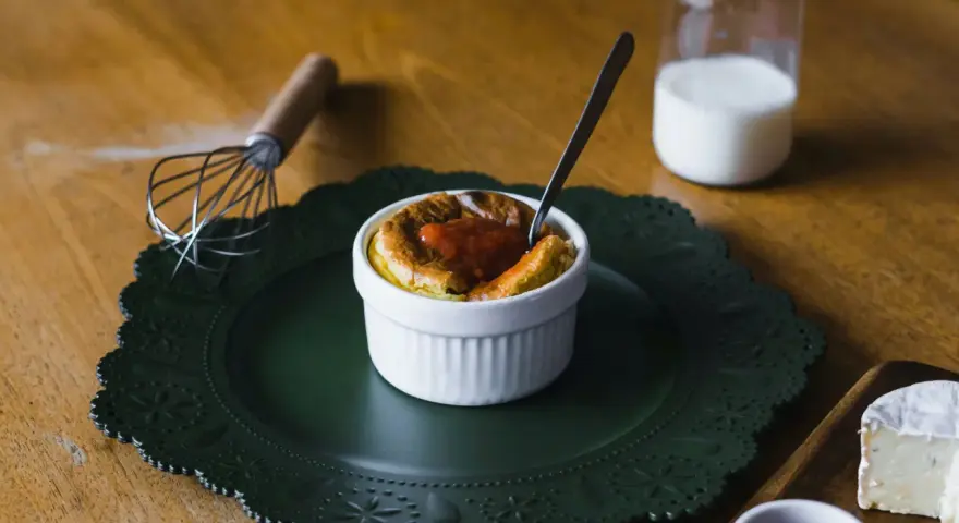 Delicioso souffle de vainilla con salsa de fresa, listo para disfrutar. A su lado, un batidor y un vaso de leche.