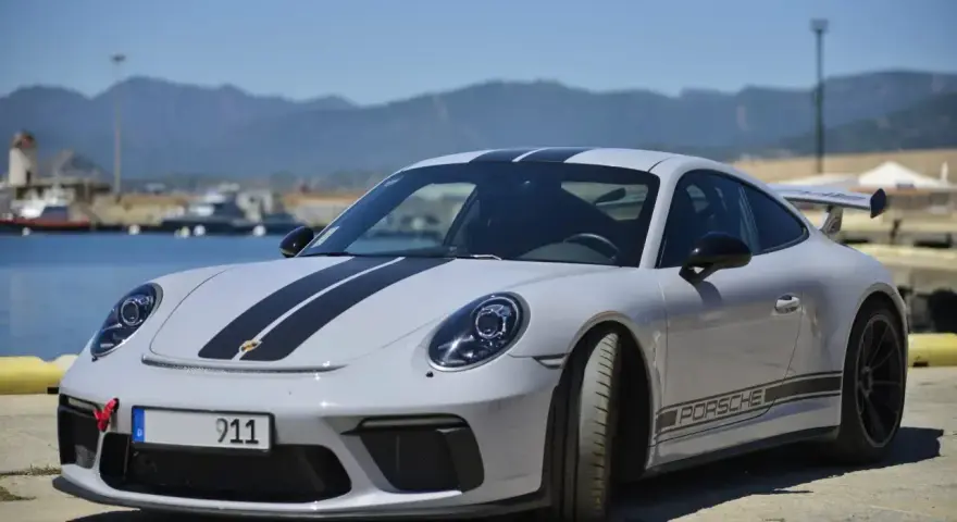Szary Porsche 911 Carrera z czarnymi pasami na masce i dachu, zaparkowany nad wodą z górami w tle.