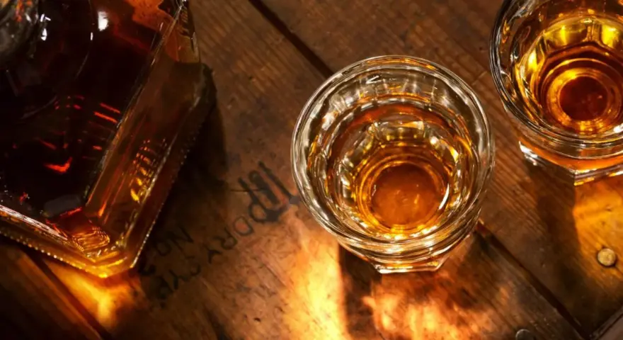 Butelka i dwa kieliszki z bursztynowym płynem, przypominającym koniak lub whisky, stoją na drewnianym blacie.