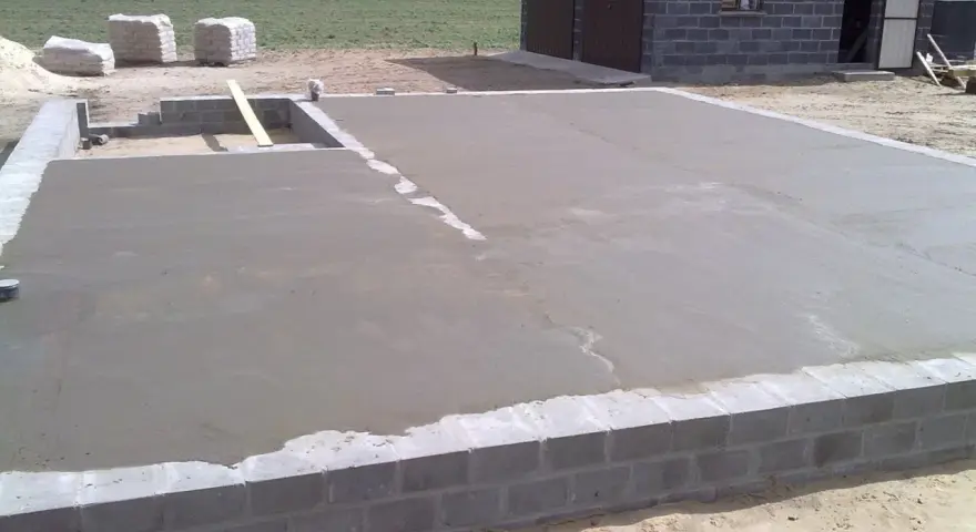 Świeżo wylany beton na chudziaka tworzy fundament pod budowę. W tle widać materiały budowlane i fragment budynku.