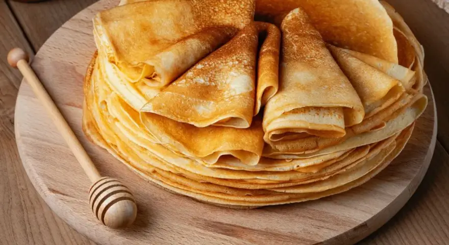 Pile de crêpes dorées, prêtes à être garnies. Une cuillère à miel en bois repose à côté, suggérant une délicieuse recette de crêpes.