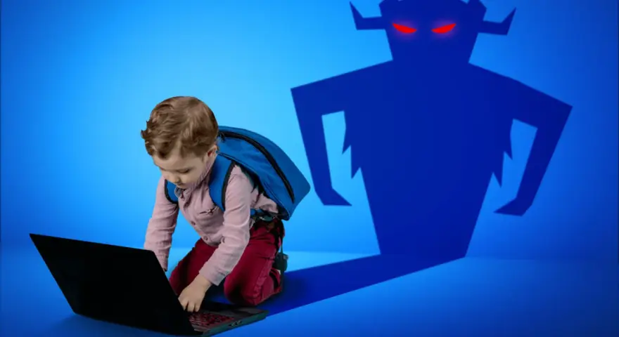Chłopiec z plecakiem pochyla się nad laptopem, a za nim cień potwora z czerwonymi oczami. Ważne jest zabezpieczenie dziecka w sieci.