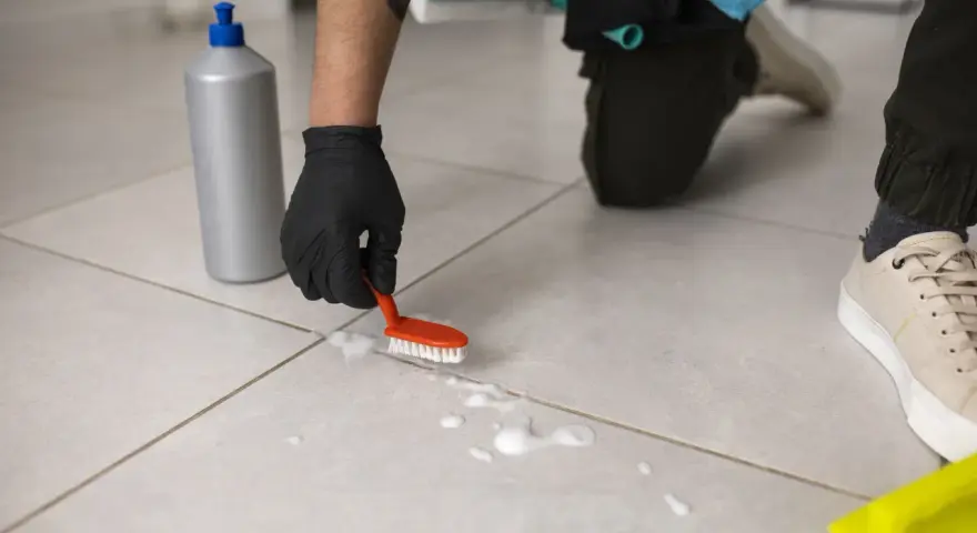 Osoba w czarnej rękawiczce czyści podłogę z płytek małą szczoteczką i pianą.