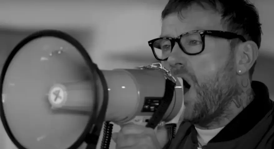 Un homme avec des lunettes et un mégaphone, sa voix résonne. Le groupe est en plein concert.