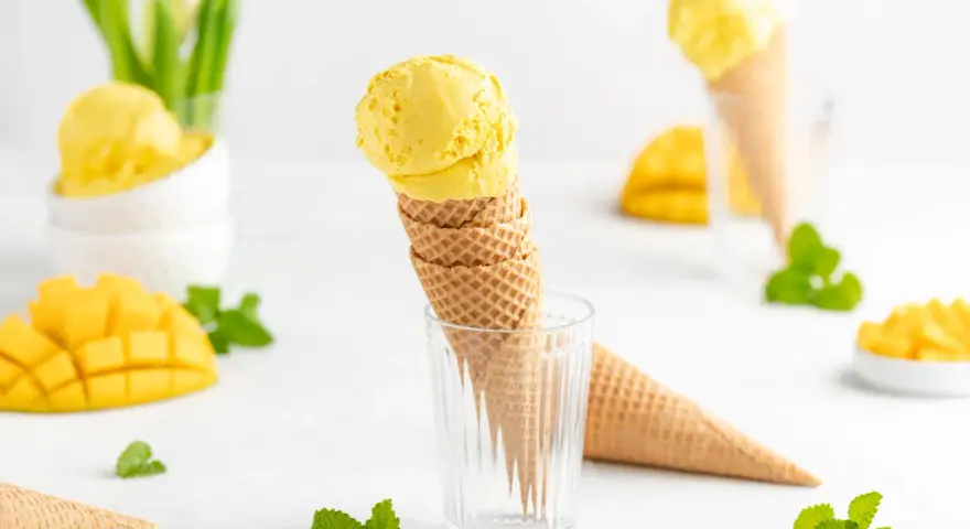 Zwei Kugeln cremiges Mango Eis in Waffelhörnchen, umgeben von frischen Mangostücken und Minze. Ein süßer Genuss!