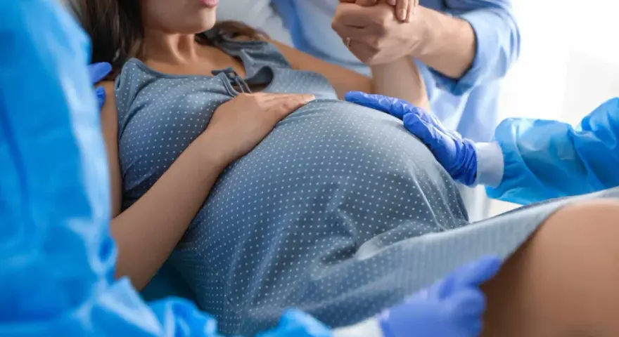 Donna in travaglio, mani che si stringono, personale sanitario con guanti blu. Si percepisce la tensione e l'attesa per lo scollamento membrane.