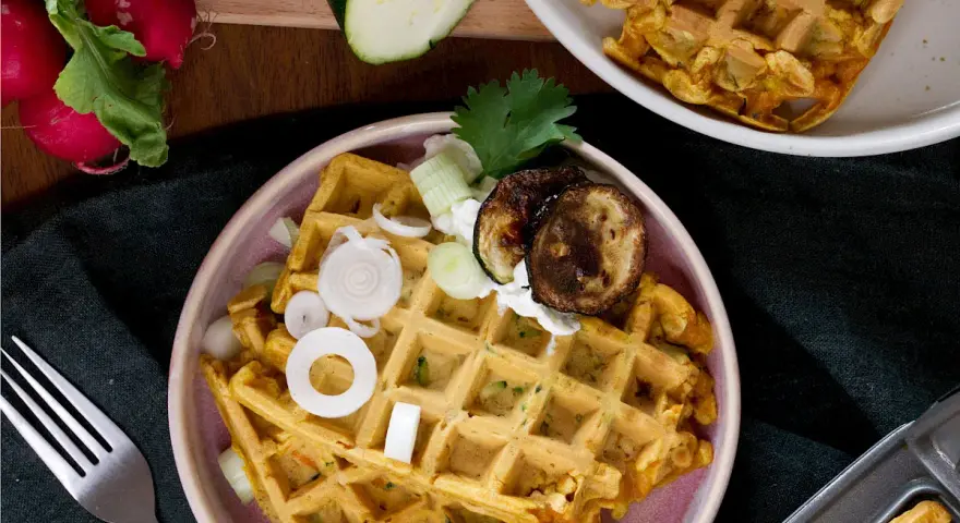 Herzhafte Waffeln mit Zucchini, Zwiebeln und gegrillten Auberginen, garniert mit Kräutern und einem Klecks Joghurt.
