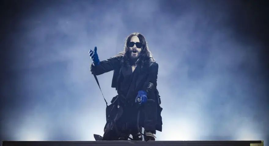 Jared Leto w niebieskich rękawiczkach, w okularach i długim płaszczu, klęczy na scenie. Jego zespół pewnie czeka na jego znak.