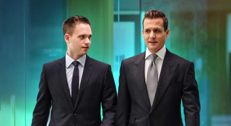 Due uomini in abiti eleganti, stile Harvey Specter, camminano in un ufficio moderno.