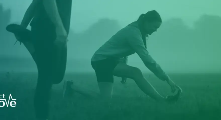 Två personer stretchar utomhus i dimma. En kvinna på knä sträcker sig mot sin fot. Detta kan vara en del av rehab benhinneinflammation.