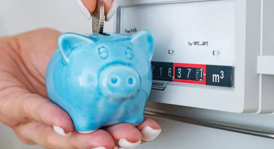 Ahorra con el plan renove calderas gas natural. Una mano deposita una moneda en una hucha cerdito azul junto a un contador de gas.