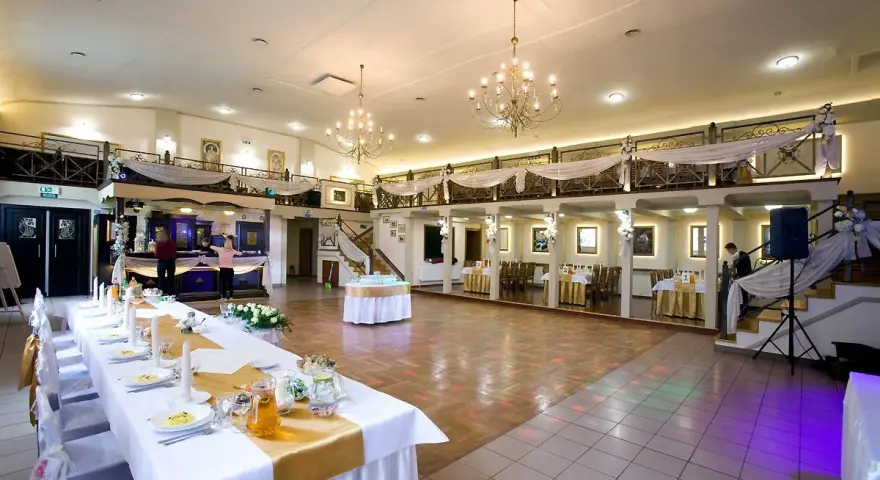 Elegancka sala weselna z długim stołem, ozdobionym złotą podkową. W tle bar i schody prowadzące na antresolę.