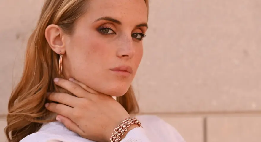 Donna con lentiggini e gioielli in oro rosa, un bracciale a maglie larghe e orecchini a cerchio.