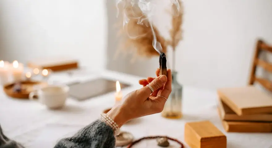 Mano sosteniendo un palo santo humeante, rodeada de velas, libros y cristales, creando un ambiente de sanación y paz.