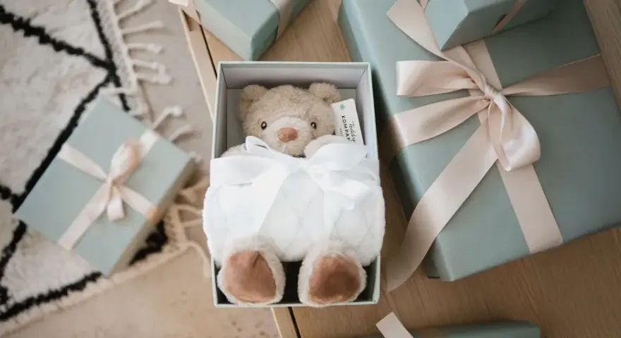 En mjuk nallebjörn i en presentbox, omgiven av fler presenter med ljusrosa band. En perfekt välkommen till världen present.