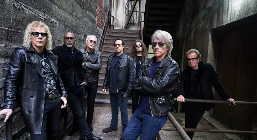 Zespół Bon Jovi gotowy na koncert, w skórzanych kurtkach i okularach przeciwsłonecznych, pozuje na tle industrialnych schodów.