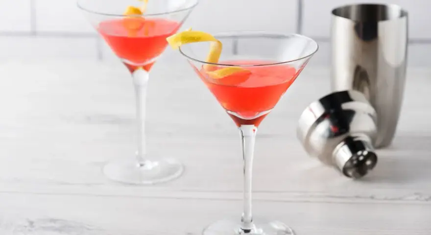 Dwa drinki Cosmopolitan z plasterkiem cytryny, obok metalowa shakera. Idealny przepis na Cosmopolitan drink.