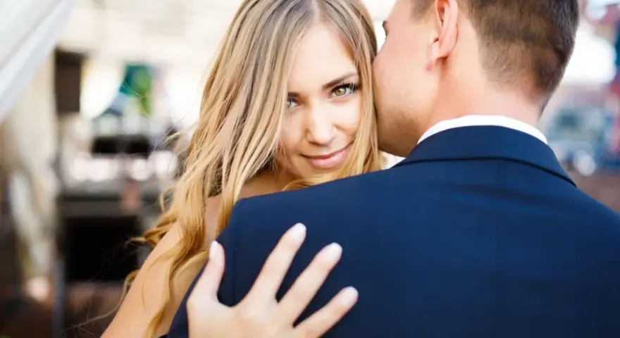 Blondynka z zielonymi oczami patrzy w obiektyw, gdy mężczyzna w granatowym garniturze, numerologiczna 8 mężczyzna, całuje ją w policzek.