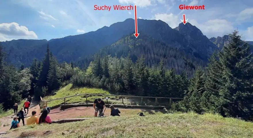Widok na góry Suchy Wierch i Giewont. Ludzie odpoczywają na szlaku, podziwiając sarnią skałę i jej wysokość.
