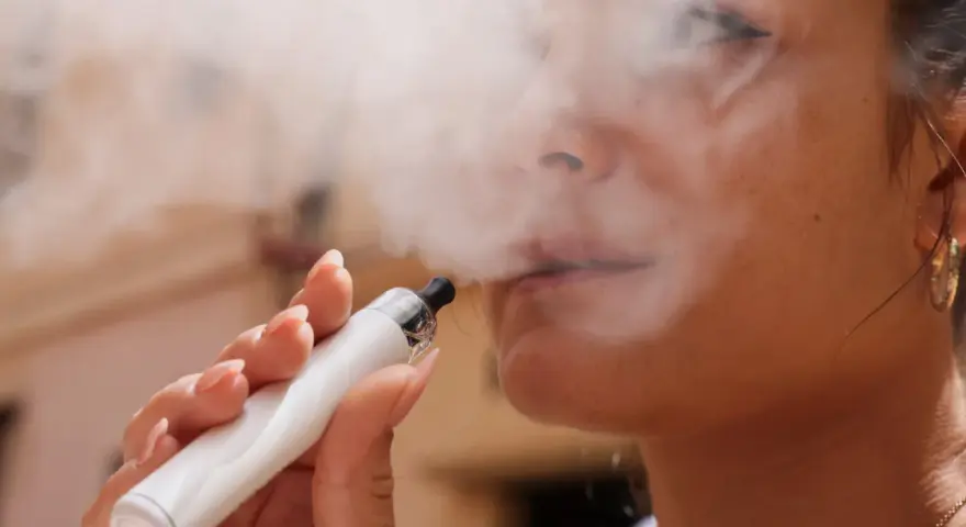 Una mujer exhala vapor de un vaper blanco. El vapor parece rodear su rostro, sugiriendo que el vaper encharca los pulmones.