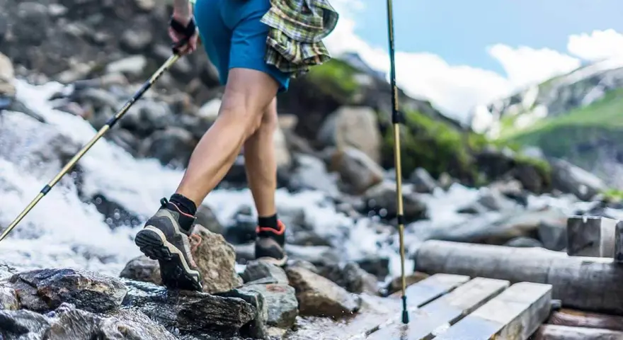 Une personne en tenue de ppg sport traverse un ruisseau sur un pont de bois, bâtons de randonnée en main. Paysage de montagne.