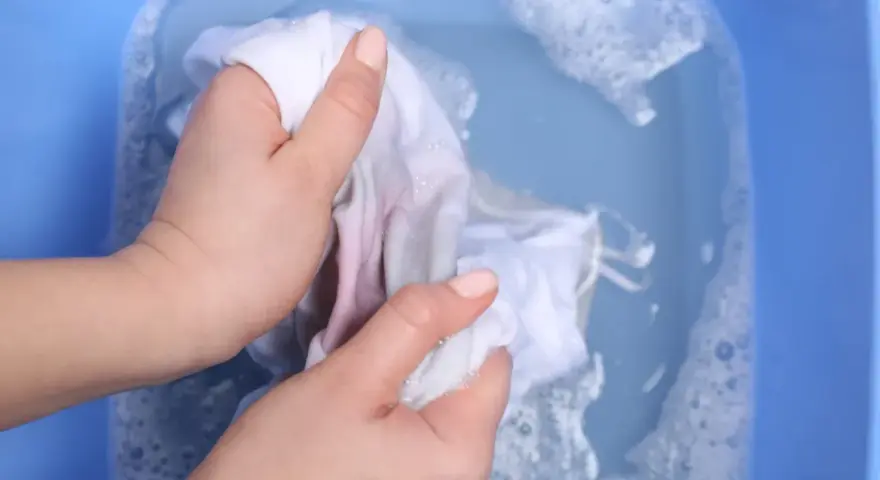 Ręce piorą białą tkaninę z delikatnymi plamami z wiśni w niebieskiej misce z wodą i pianą.