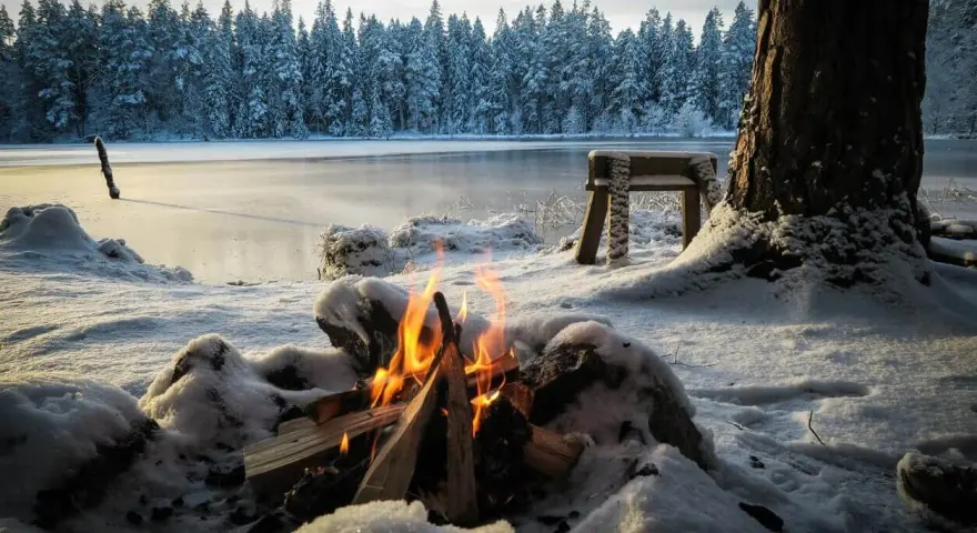 Ein Lagerfeuer wärmt beim Zelten im Winter am gefrorenen See. Schnee bedeckt die Ufer und den Wald im Hintergrund.