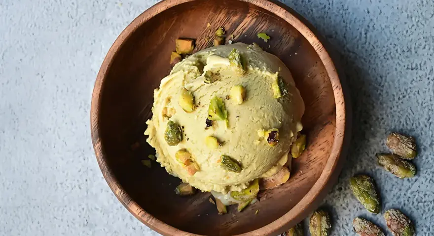 Cremoso helado de pistacho servido en un cuenco de madera, adornado con trozos de pistacho y acompañado de más pistachos enteros esparcidos alrededor.